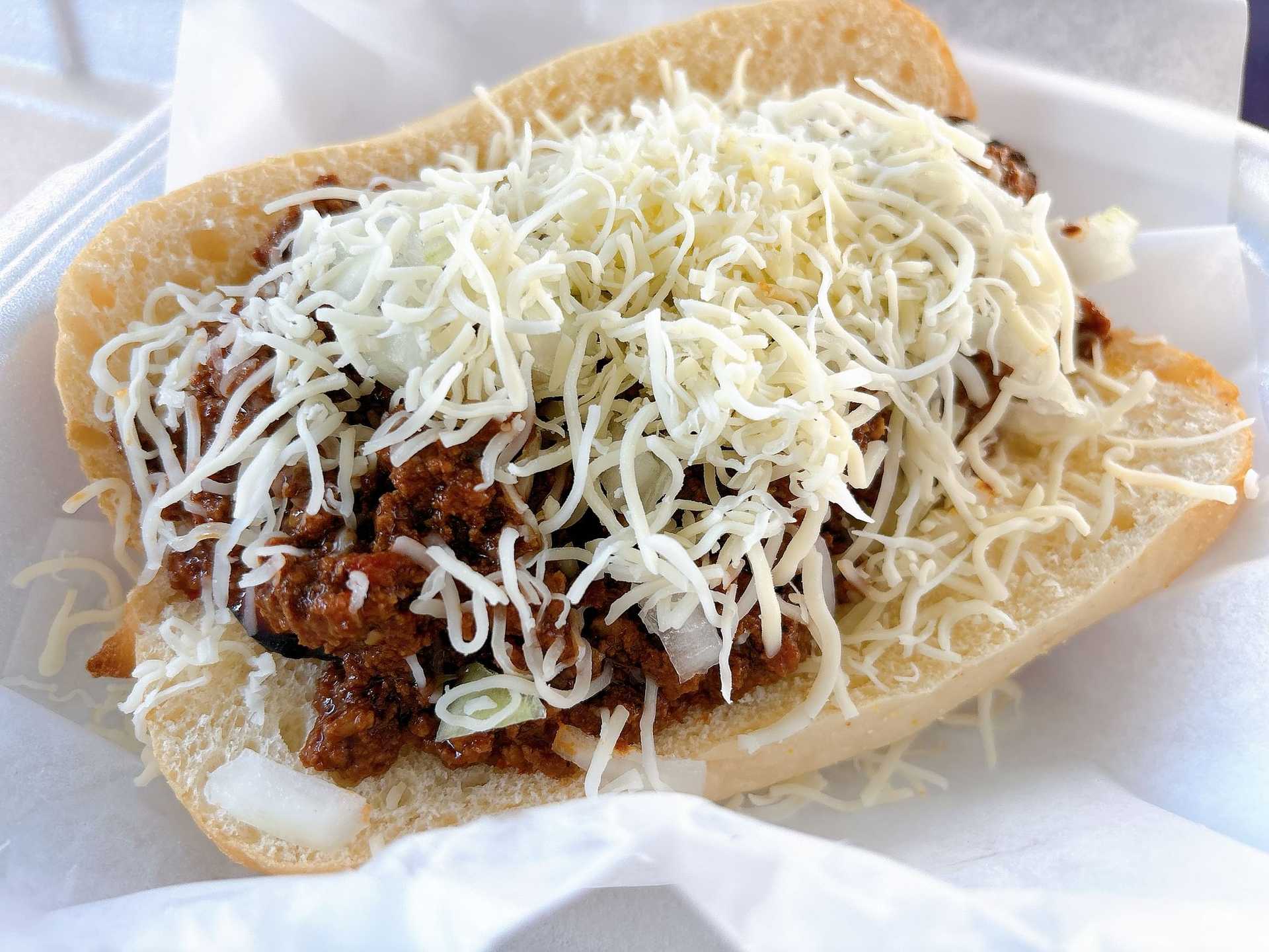 Chili cheese dog topped with shredded cheese and onions in a soft bun on a paper-lined plate.